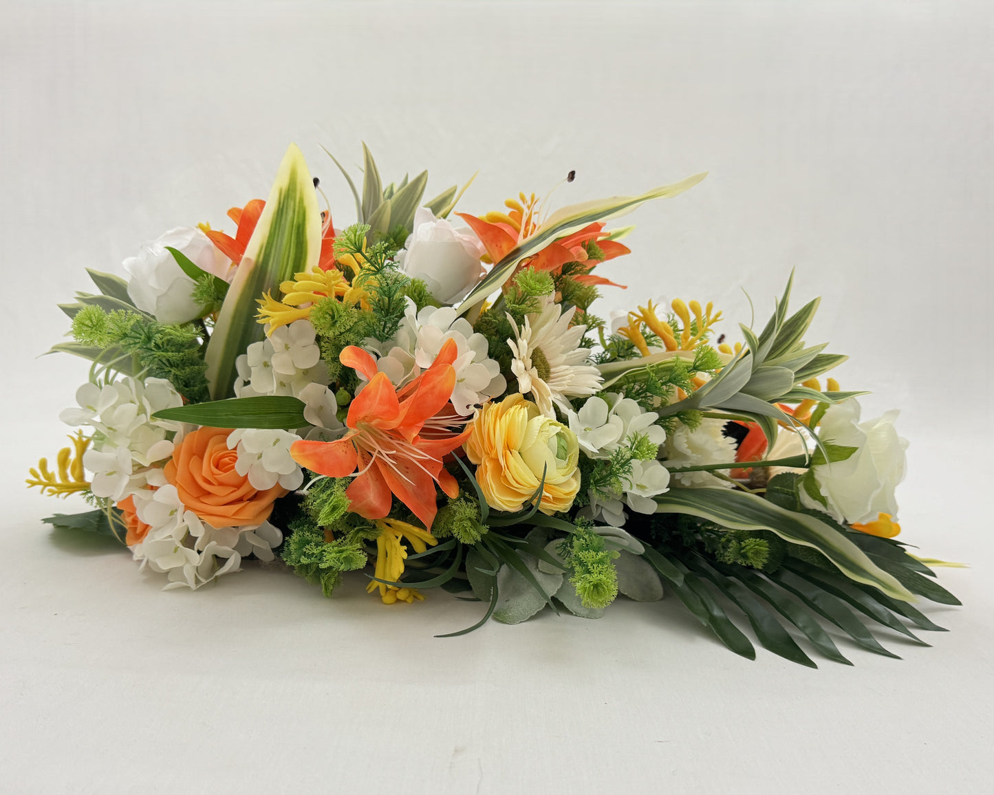 Druppelvormig rouwstuk met oranje en gele tinten. Zonnebloemen, lelies en rozen. Van de zijkant gezien.
